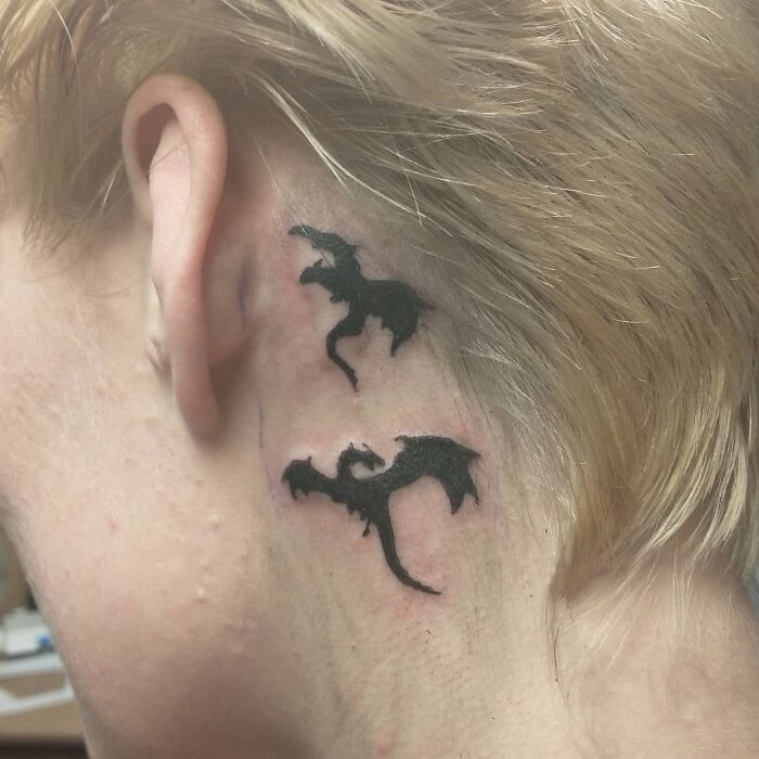 Ear tattoo of two black dragons behind the ear against blonde hair.