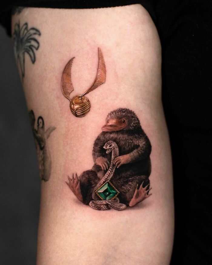 Harry Potter tattoo of a Niffler holding a snake with an emerald, beneath a Golden Snitch, on the upper arm.
