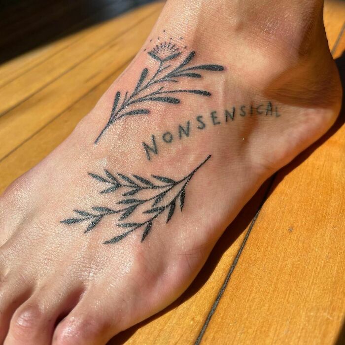 Word tattoo with "nonsensical" and floral design on foot, sunlight casting shadows on wooden floor.
