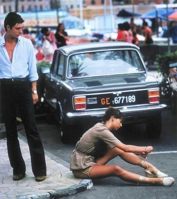 Un hombre se detiene para ver a una mujer atarse las sandalias en el puerto deportivo de Portofino, agosto de 1977. Foto de Slim Aarons