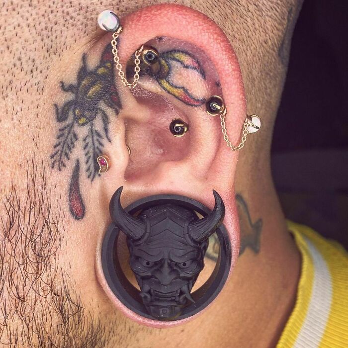 Ear tattoo with bold design, featuring piercings and a large black plug with a demon face.