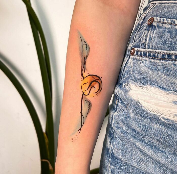 Harry Potter tattoo of the Golden Snitch on a person's forearm, wearing ripped jeans, against a leafy background.