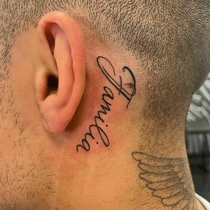 Ear tattoo of the word "familie" in elegant script behind the ear.