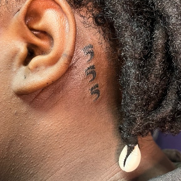 Ear tattoo featuring black symbols next to locks of hair adorned with a white bead.