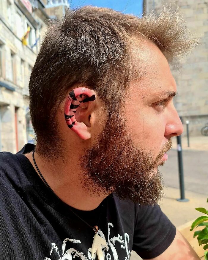 A man with a unique ear tattoo featuring black and pink tiger stripes stands outside wearing a black shirt.