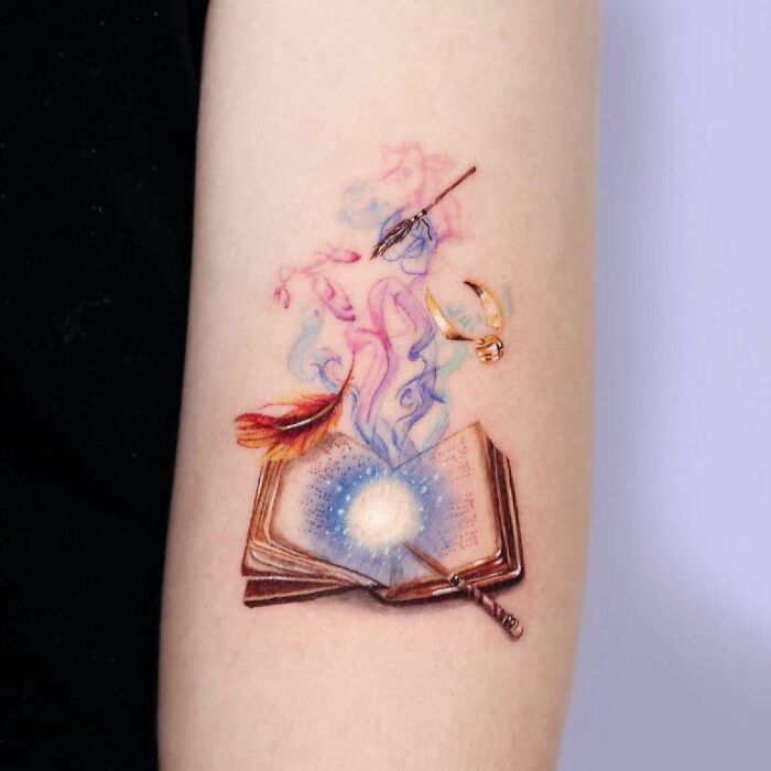 Colorful tattoo of an open book with a wand, quill, and Golden Snitch, inspired by the wizarding world.