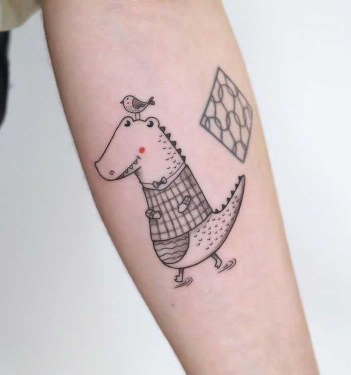 Cartoon dinosaur tattoo with bird on arm, showcasing cute animal tattoo art.