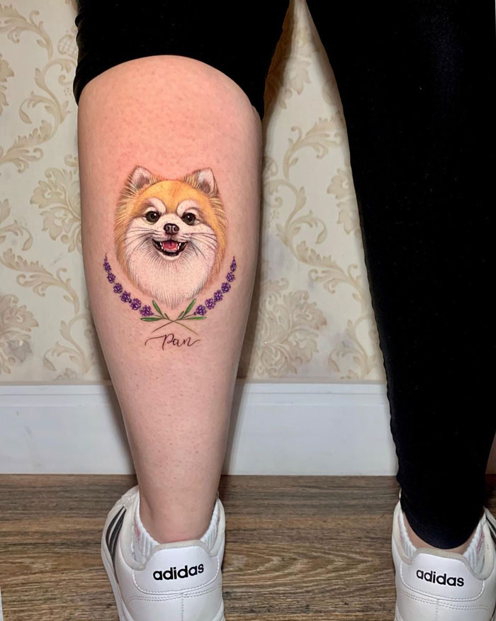 Unique calf tattoo of a smiling dog with floral accents, worn by a person in black pants and white Adidas sneakers.