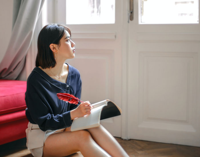 A young woman sitting on the floor, holding a notebook and red feather