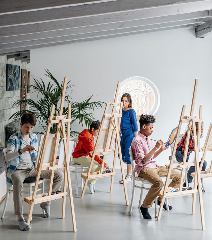 Woman teacher walks between children while they sit near their easels and paint in art class