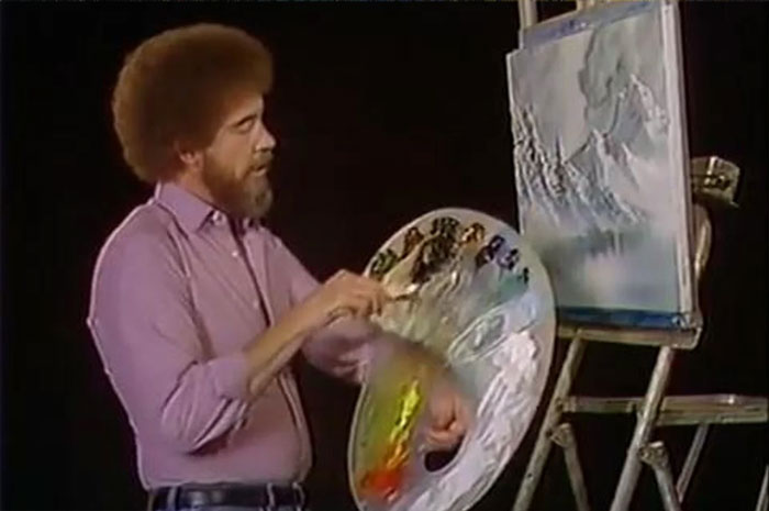 Bob Ross in pink shirt holds a palette and paints a picture in gray