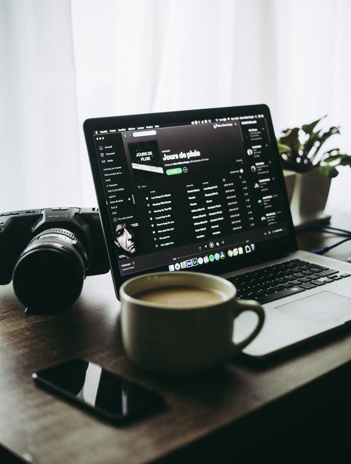 A laptop with playlist on it next to camera, phone and cup of coffee 