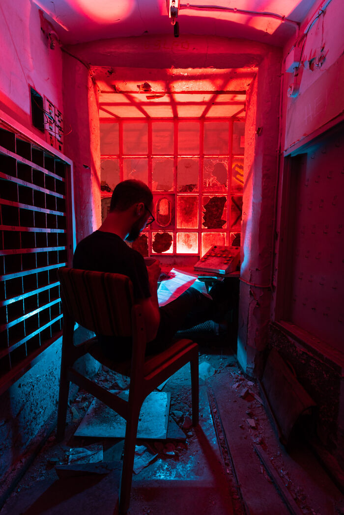 Man sitting in an abandoned place at night illuminated by vibrant red and blue light, reading papers in a dark, rundown room.