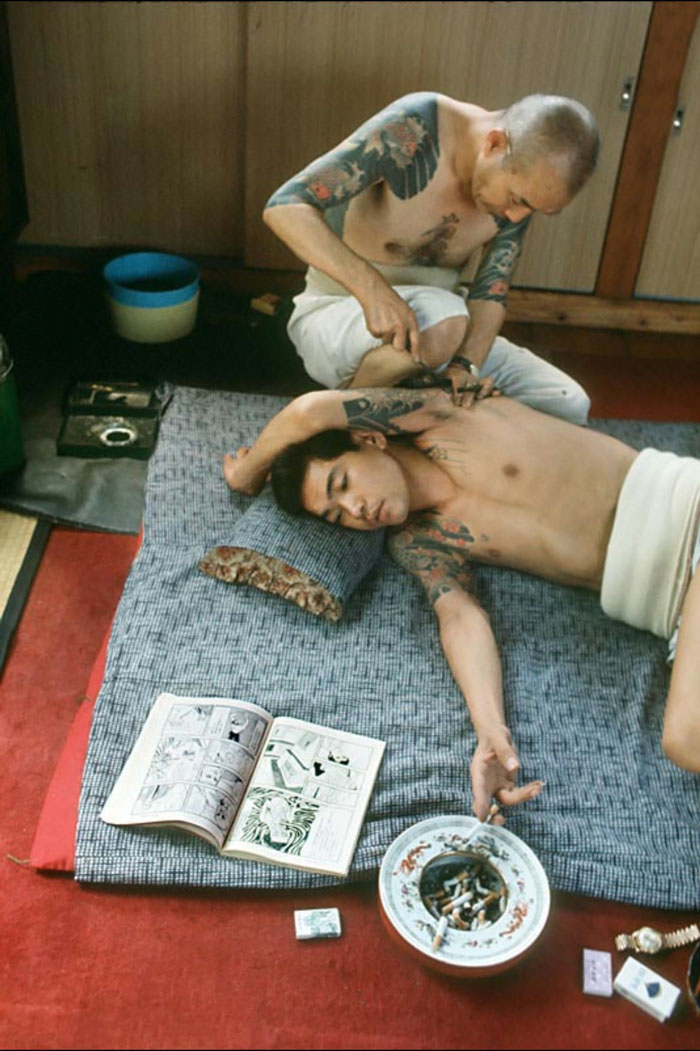 Martha Cooper, Hombre fumando un cigarrillo y leyendo un cómic mientras le tatúan, Tokio, Japón, 1970