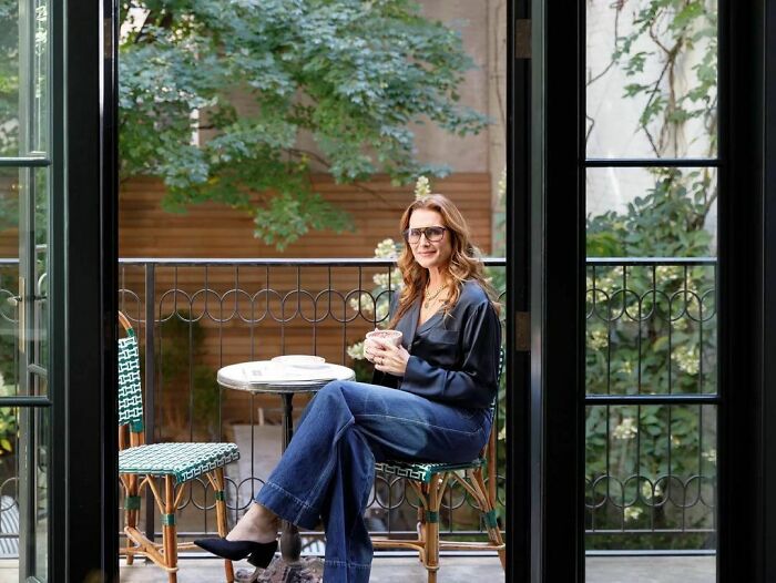 Woman wearing glasses and dark blouse sitting on a balcony chair, holding a cup, representing intelligent celebrities.