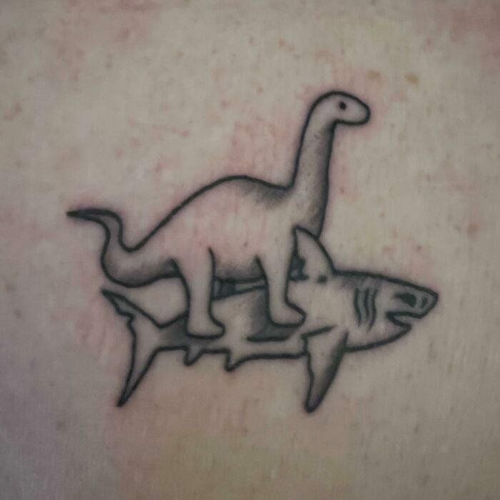 Funny tattoo of a dinosaur riding a shark on skin.