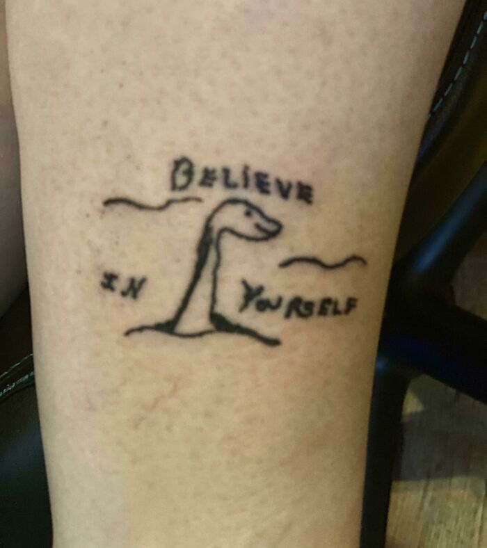 Tattoo of a doodle dinosaur with the humorous phrase "Believe in Yourself" on skin.