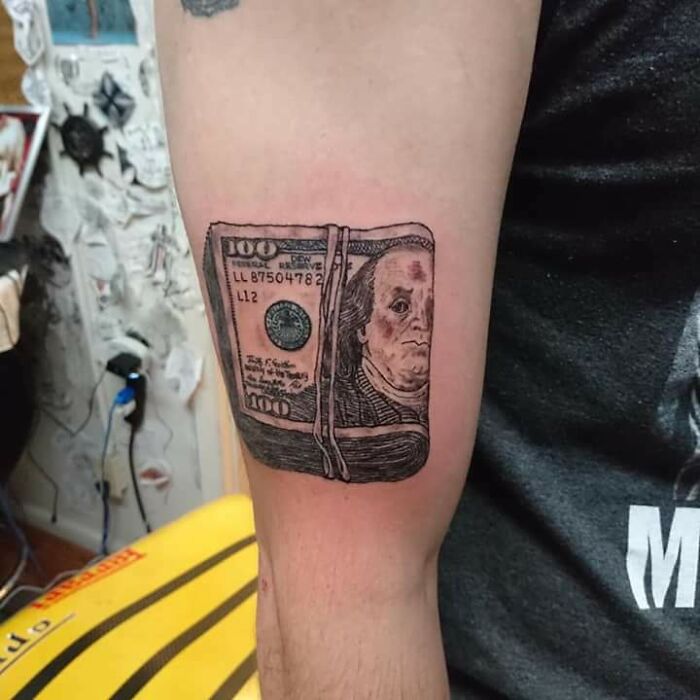 Funny tattoo of sad Ben Franklin on arm, humorously depicting a bundle of $100 bills.