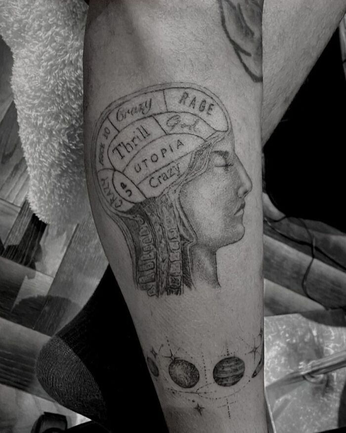 Celebrity tattoo featuring a phrenology head with words and planetary symbols on a leg.