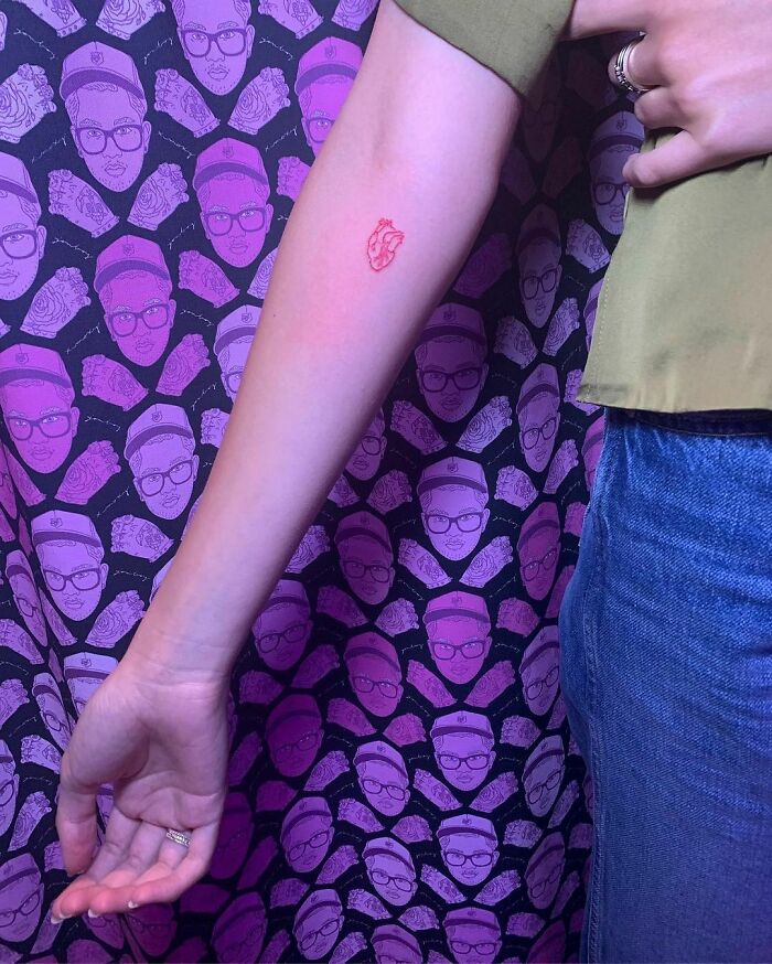 Arm displaying a small red heart tattoo against a purple patterned background with celebrity faces.