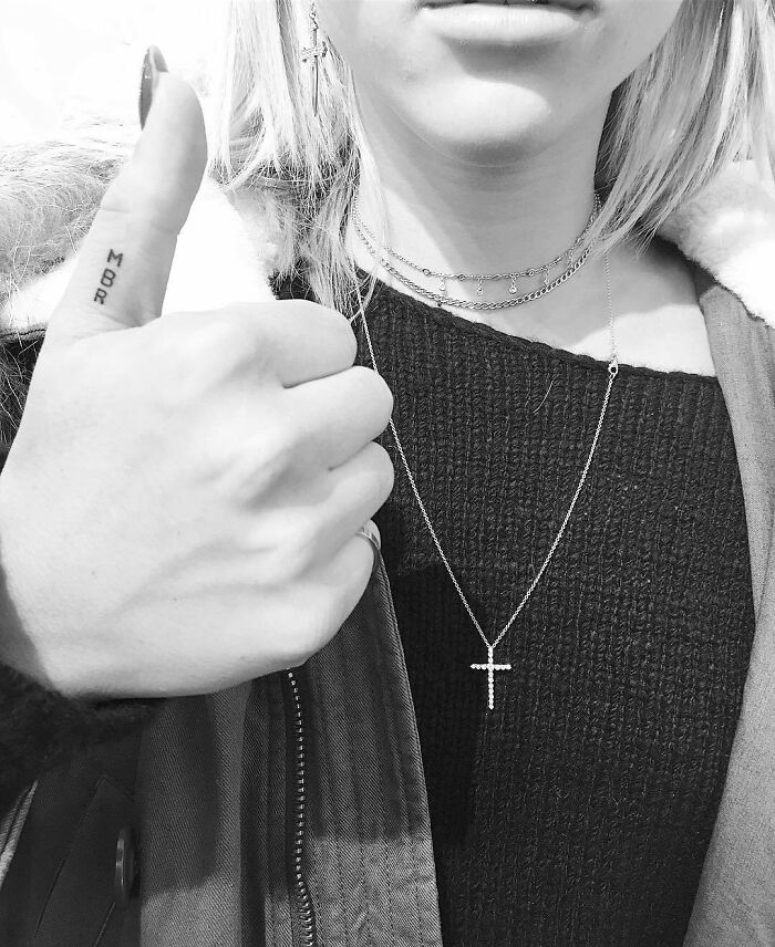 Woman shows a tattoo on her thumb, wearing a cross necklace and a cozy jacket.