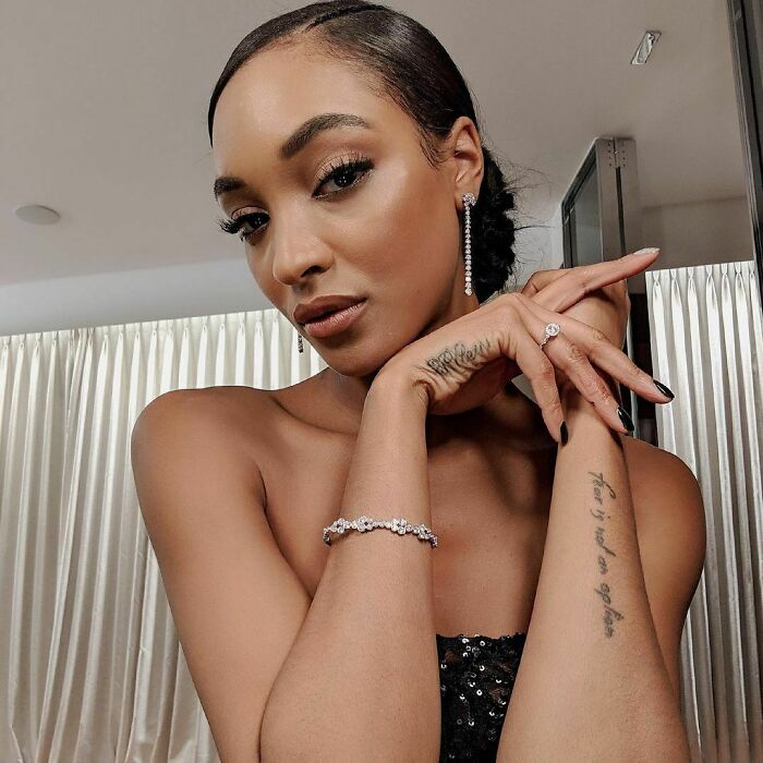 Celebrity with arm tattoo, wearing diamond jewelry, posing elegantly in a chic setting.