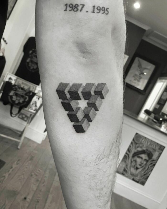 Optical illusion tattoo of a 3D impossible triangle on forearm, with text "1987 . 1995" above.
