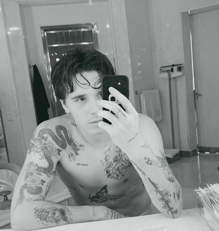 Man with celebrity tattoos takes a mirror selfie in a bathroom.