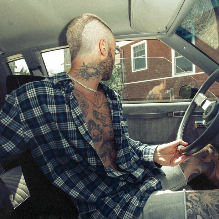 Man driving a car, showing off star tattoos on his neck and chest, wearing a plaid shirt and shorts.