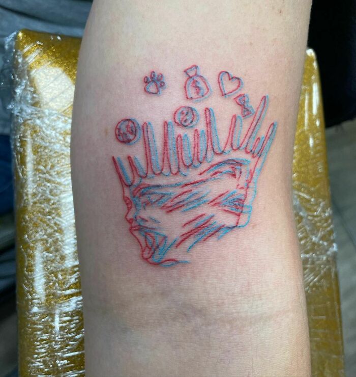 Optical illusion tattoo in red and blue ink, featuring a distorted crown and face design on a forearm.