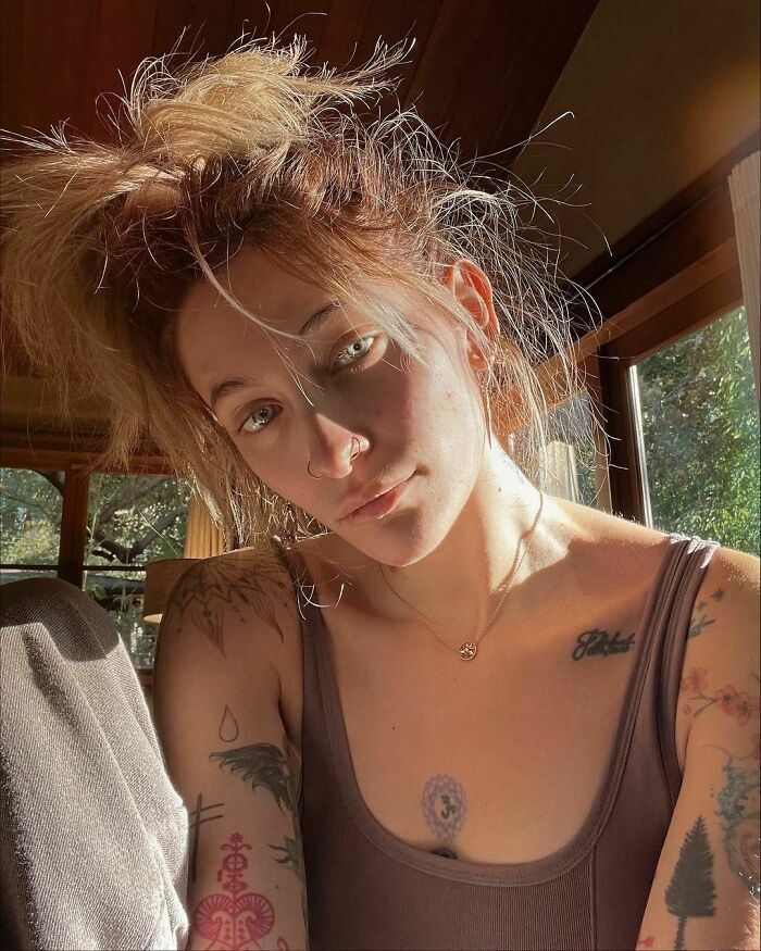 Person with various tattoos on arms and chest, wearing a tank top, sitting by a sunlit window.