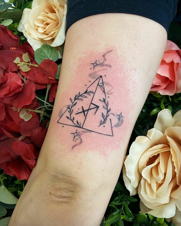 Arm tattoo of the Deathly Hallows from Harry Potter, surrounded by vines and stars, set against red and white roses.
