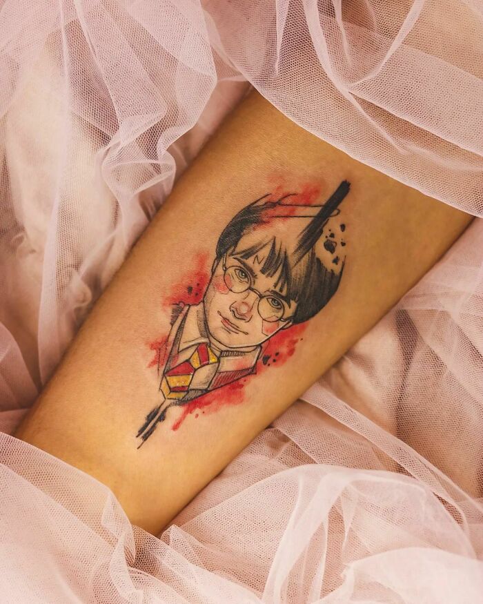 Harry Potter tattoo on arm, featuring character's face and glasses, surrounded by red and black artistic splashes.