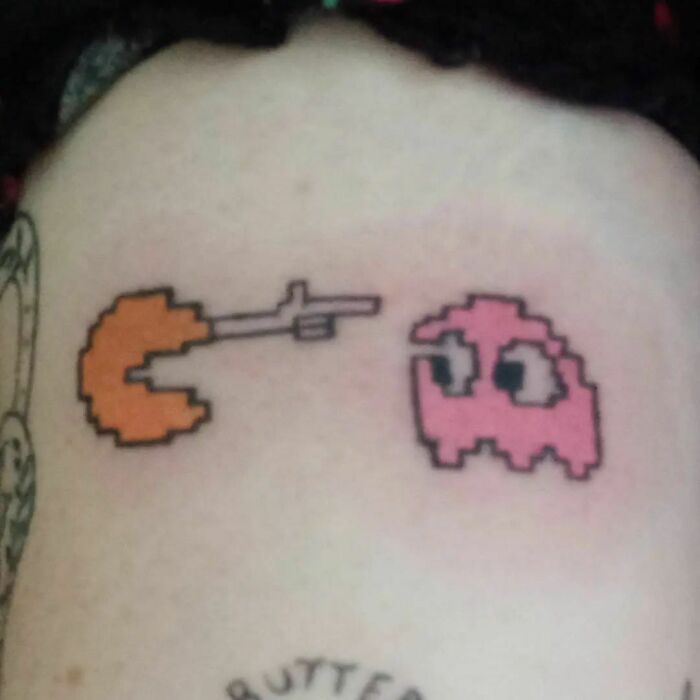 Funny tattoo of a pixelated orange Pac-Man with a gun chasing a pink ghost on skin.