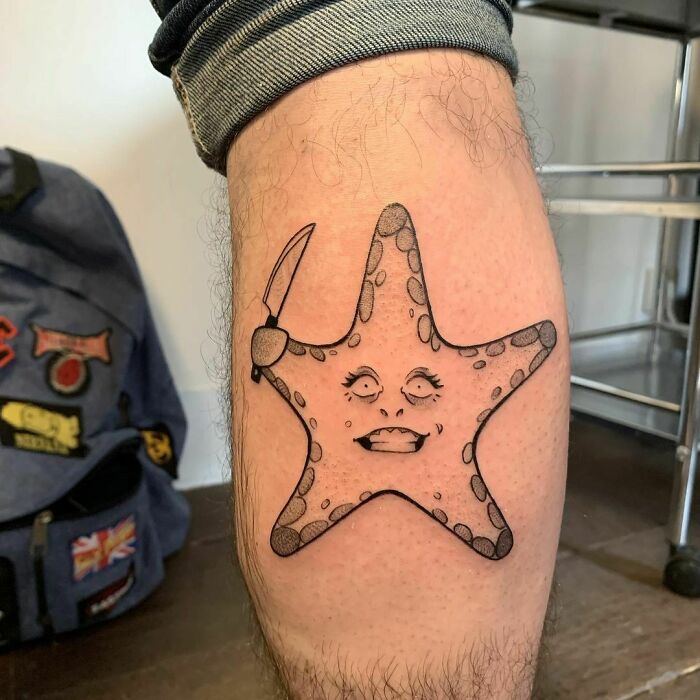 Funny tattoo of a starfish with a knife leg, featuring a quirky face, on a person's leg.