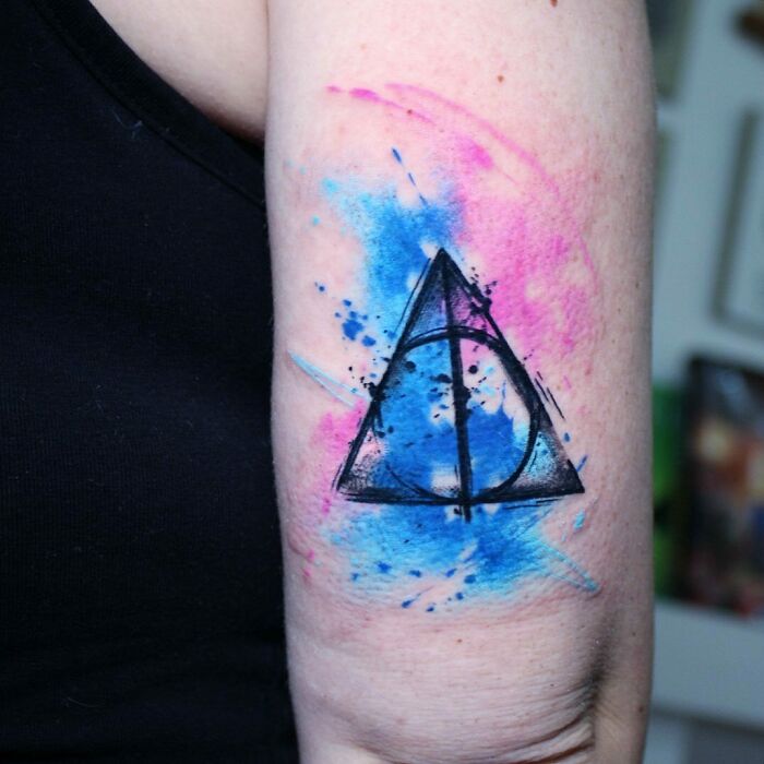 Harry Potter tattoo featuring the Deathly Hallows with blue and pink watercolor design on upper arm.