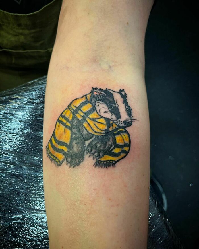 Badger tattoo with a yellow and black scarf, symbolizing Harry Potter's Hufflepuff house, on a forearm.