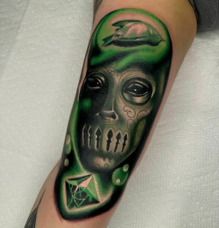 Green and black Harry Potter tattoo on forearm featuring a mystical face and a gem.