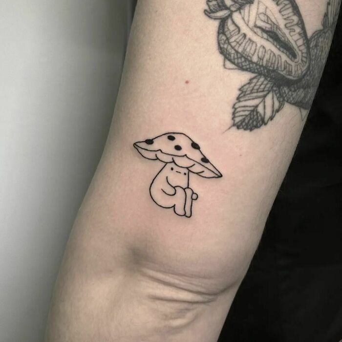 Whimsical tattoo of a cute character sitting under a mushroom, showcasing funny tattoo art on an arm.
