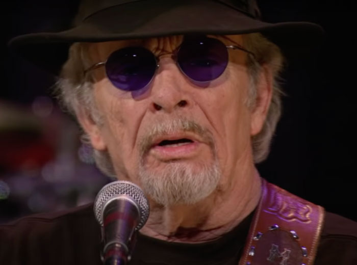Merle Haggard wearing glasses and hat singing 