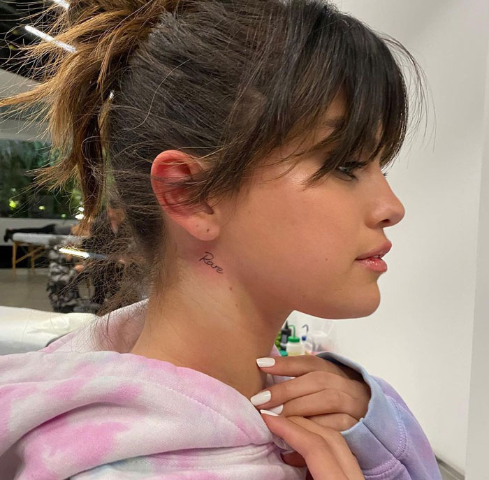 Selena Gomez with a tattoo behind the ear, wearing a pastel hoodie and looking sideways.