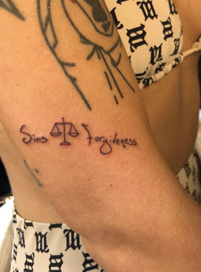 Celebrity tattoos featuring "Sins Forgiveness" phrase on arm with scales symbol.