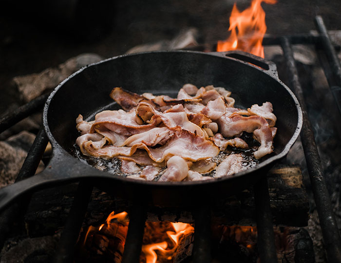 Cast iron skillet frying bacon over an open flame, showcasing outdoor cooking and big trends in rustic cooking styles.
