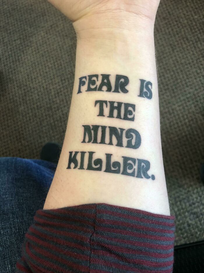 Arm tattoo reading "Fear is the mind killer" in bold black font on pale skin.