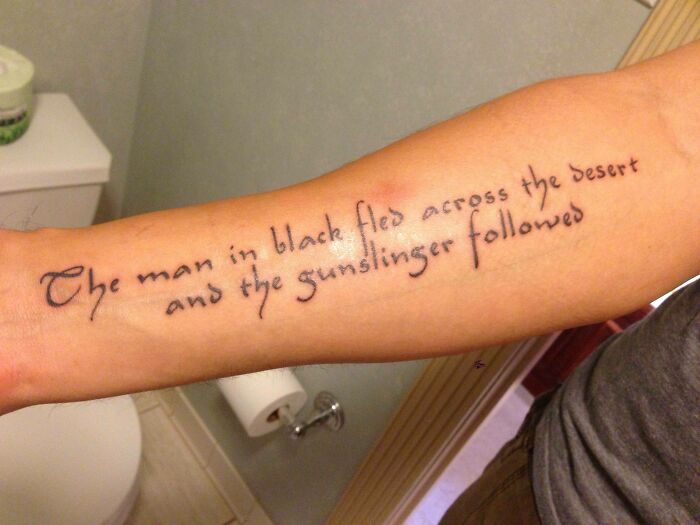 Tattooed arm with the phrase, "The man in black fled across the desert and the gunslinger followed," in cursive writing.