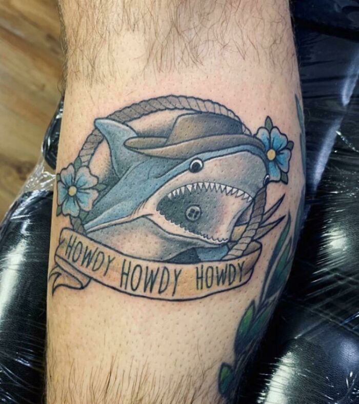 Shark tattoo with cowboy hat and “Howdy Howdy Howdy” banner, featuring blue flowers, on skin.