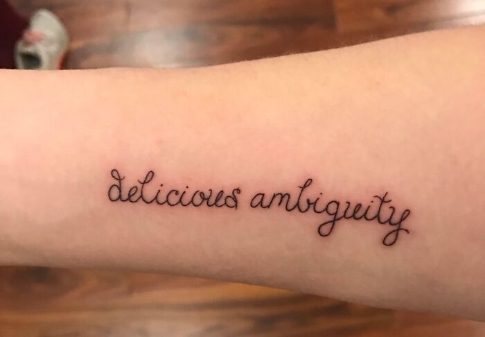 Tattoo with the words "delicious ambiguity" inked on an arm, showcasing elegant script.