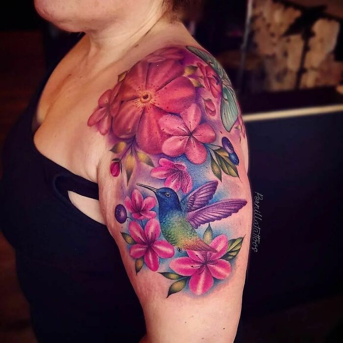 Colorful nature tattoo featuring a hummingbird and pink flowers on a woman's shoulder.