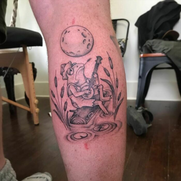 Nature tattoo of a frog playing a banjo under a moon, surrounded by cattails on a person's leg.