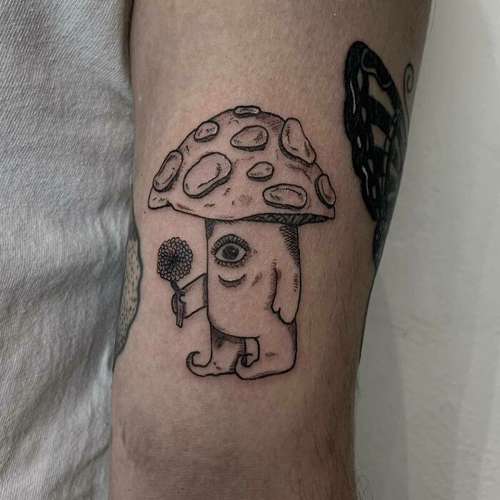Nature tattoo of a whimsical mushroom character on an arm, featuring an eye and holding a small object.